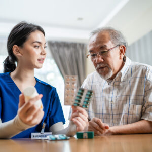 Asian Senior elderly male patient consult with physician nurse at nursing home care. Caregiver therapist pharmacist girl hold medicine pills bottle, explain prescription to older disabled guy in house